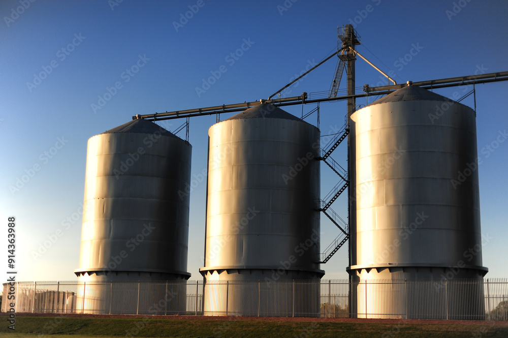 A lateral low-angle view of three steel and chrome industrial grain silos A lateral low-angle view of three steel and chrome industrial grain silos