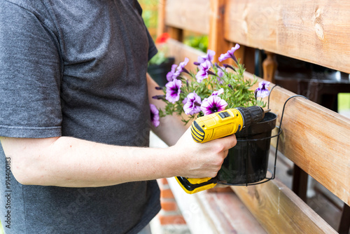 A person uses a cordless drill to attach a hanging planter full of purple flowers to a wooden fence.