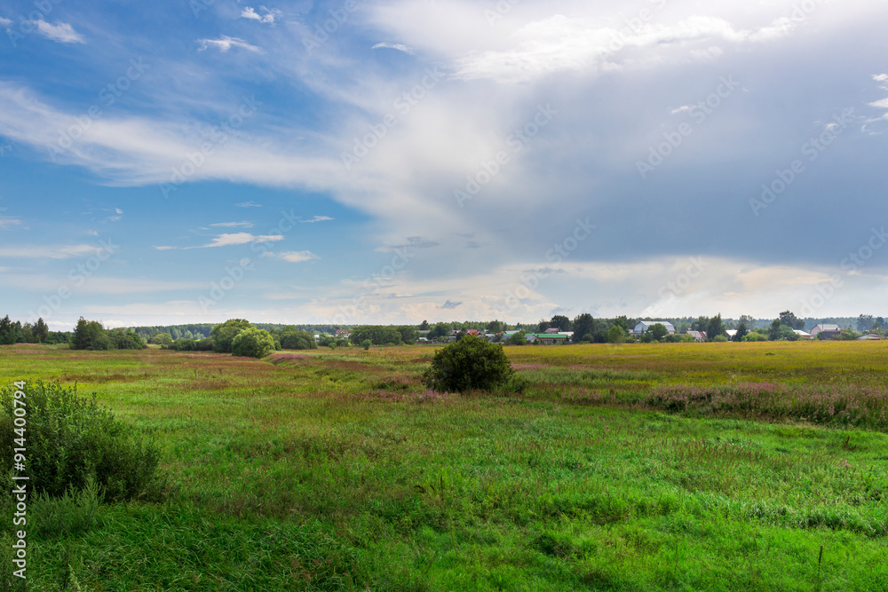 A lush green meadow stretches under a vibrant blue sky, dotted with clouds, offering a serene view of nature and a quaint rural village in the background. A lush green meadow stretches under a vibrant blue sky, dotted with clouds, offering a serene view of nature and a quaint rural village in the background.