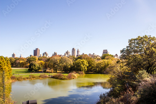 Sunny day view of a Central Park lake surrounded by trees, with a backdrop of skyscrapers in the distance