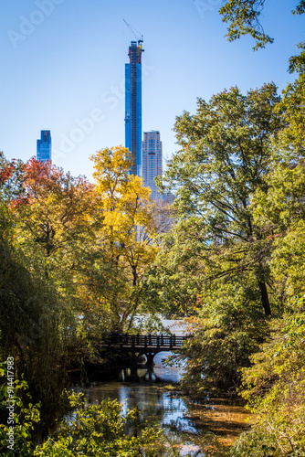 Photograph of Central Park in autumn, featuring trees, a lake, and a wooden bridge, with new skyscrapers in New York City under construction in the background on a sunny day