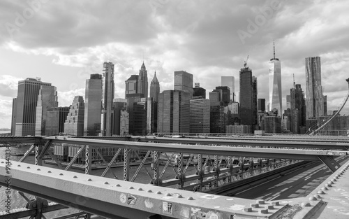 Black and white close-up of the industrial steel beams of the Brooklyn Bridge, with Manhattan's skyscrapers and the Hudson River in the background. A high-contrast urban and industrial scene