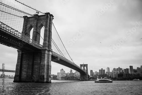 Black and white photograph of the Brooklyn Bridge, the Hudson River, and the Brooklyn neighborhood in the background on a cloudy day, with a small yacht in view.