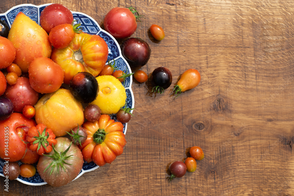 Many garden tomatoes in different colors, shapes and varieties on a blue rustic plate on a wooden table. Tomatoe composition with space for text.