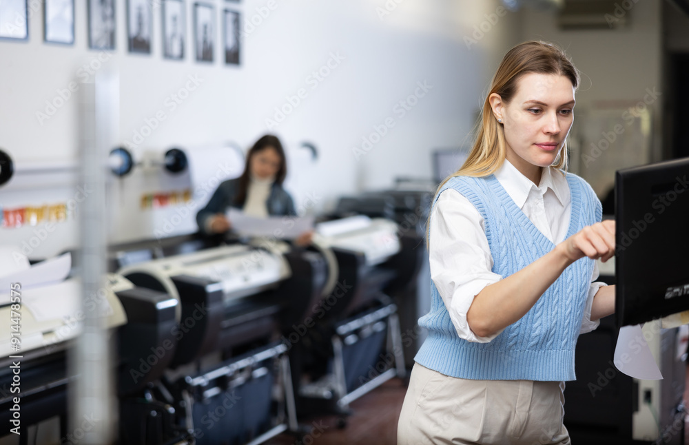 Fototapeta premium Woman using printer while working in print shop. Female printshop worker using printing device, pushing buttons.