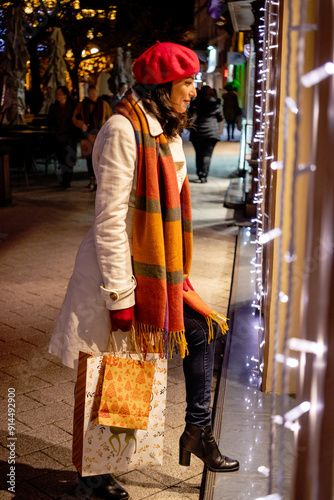 A woman in a coat and a red French beret with shopping bags enjoys cheerful Christmas shopping standing in front of a store window.