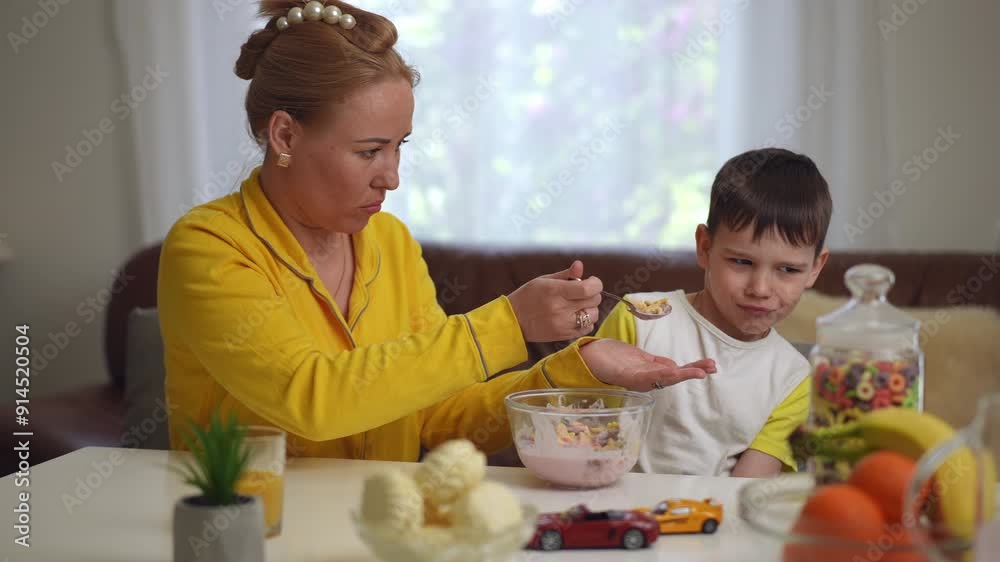 A mother holds a tablespoon to her small son's mouth, trying to feed him a breakfast cereal with yogurt, while sitting with him at the dining table in the living room. The child refuses to eat