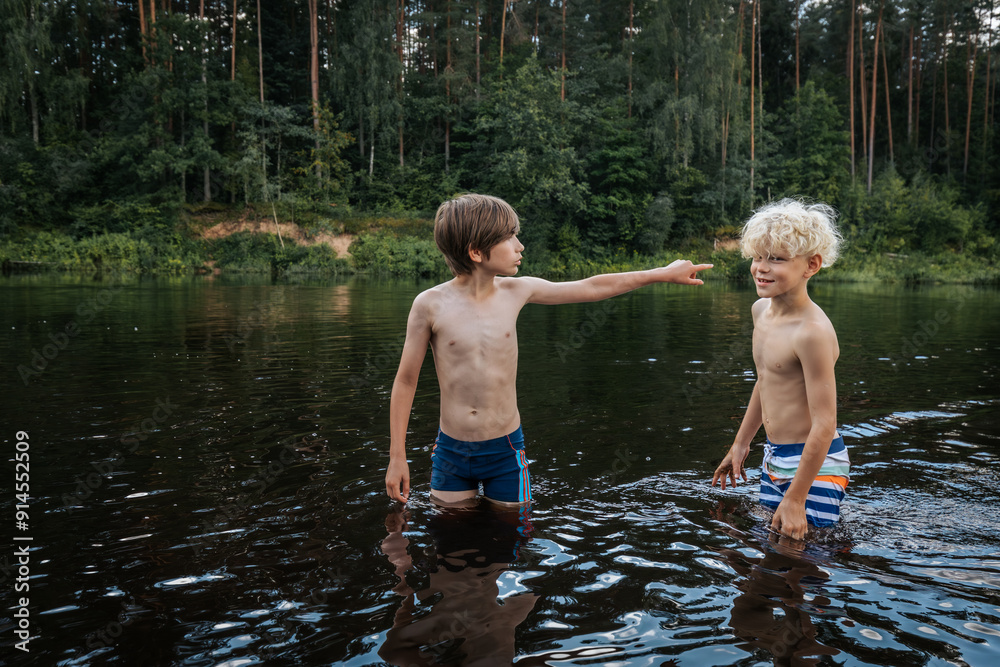 Fototapeta premium Two young boys stand in a calm river, surrounded by lush green forest. One boy points something out to the other, as they both enjoy the refreshing water. The image captures a moment of childhood