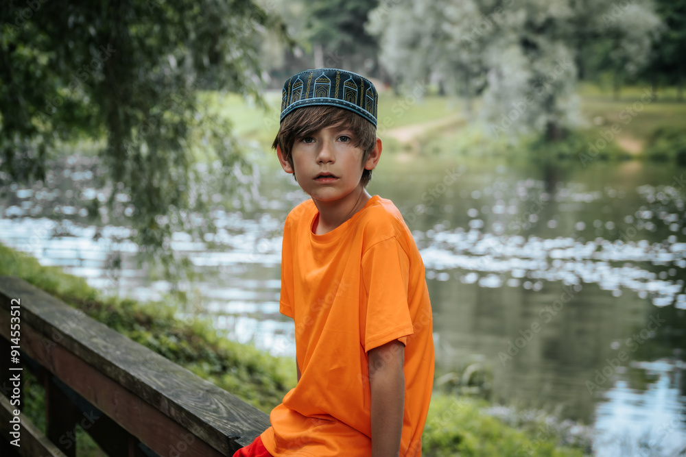 A young boy wearing an orange shirt and red shorts sits on a wooden bridge in a park. The lush greenery and a calm river in the background create a peaceful outdoor scene A young boy wearing an orange shirt and red shorts sits on a wooden bridge in a park. The lush greenery and a calm river in the background create a peaceful outdoor scene