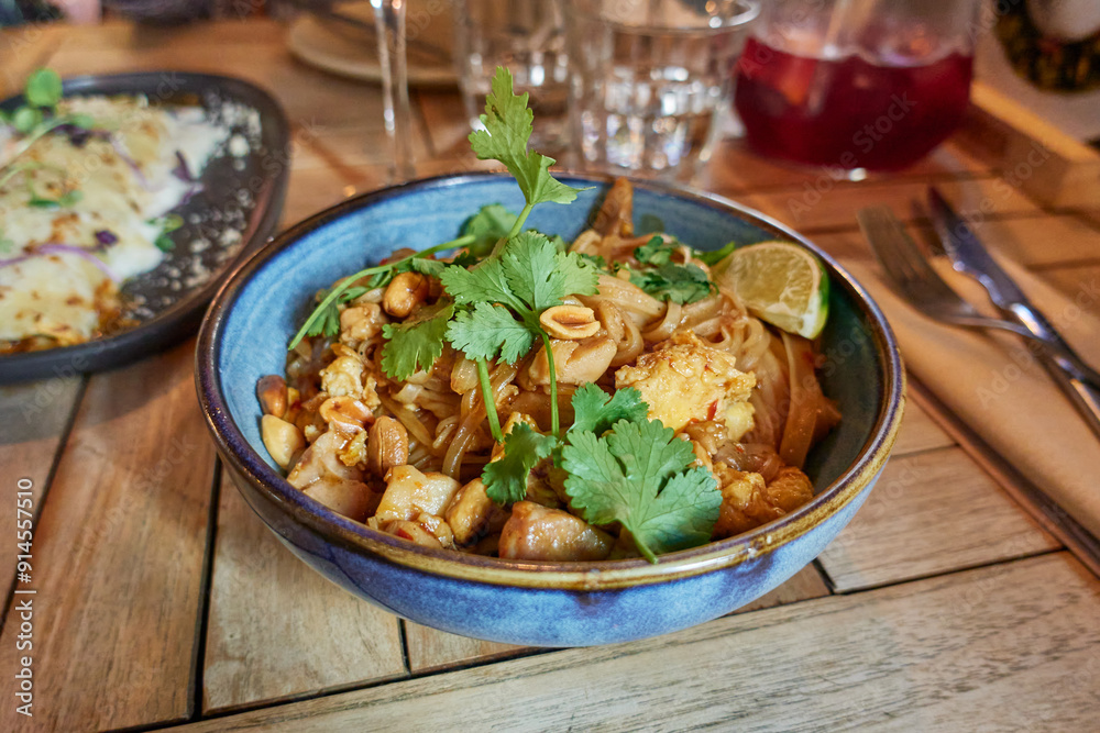 Pad Thai with chicken, peanuts, egg, lime sliced and green cilantro leaves in a blue bowl on a restaurant wooden table or at home
Pad Thai with chicken, peanuts, egg, lime sliced and green cilantro leaves in a blue bowl on a restaurant wooden table or at home