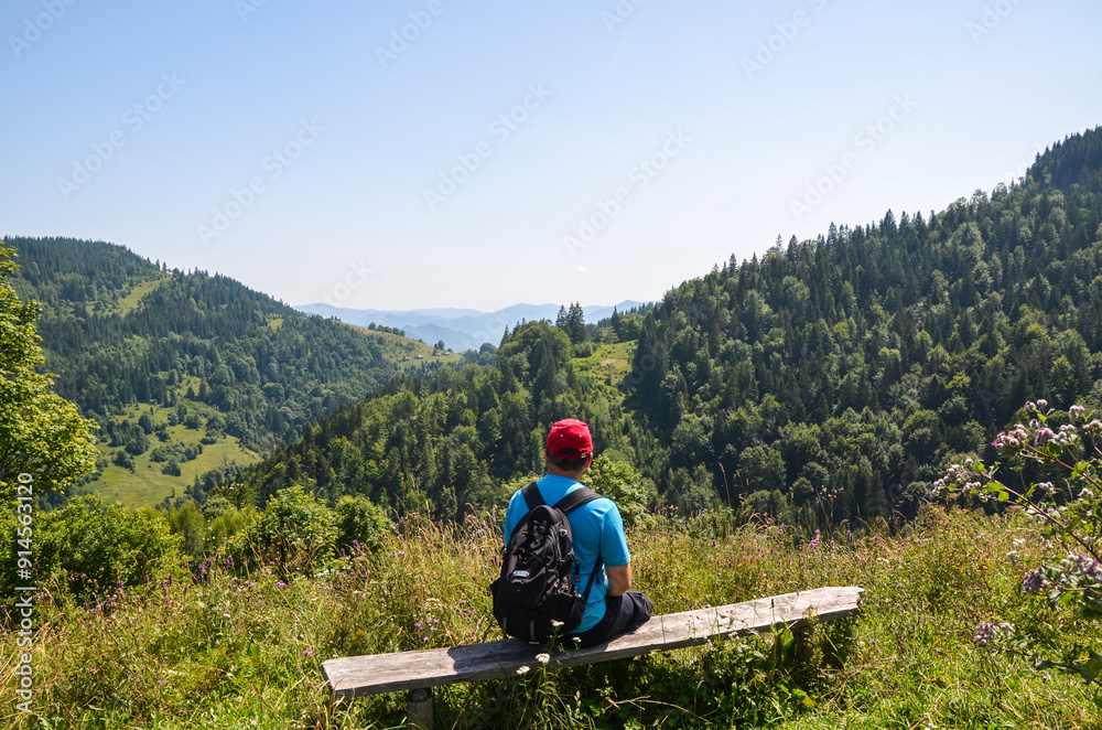 Man hiker with backpack sitting on a wooden bench, facing away from the camera, looking out over a lush green valley with rolling hills, mountains and dense forests under a clear blue sky Man hiker with backpack sitting on a wooden bench, facing away from the camera, looking out over a lush green valley with rolling hills, mountains and dense forests under a clear blue sky