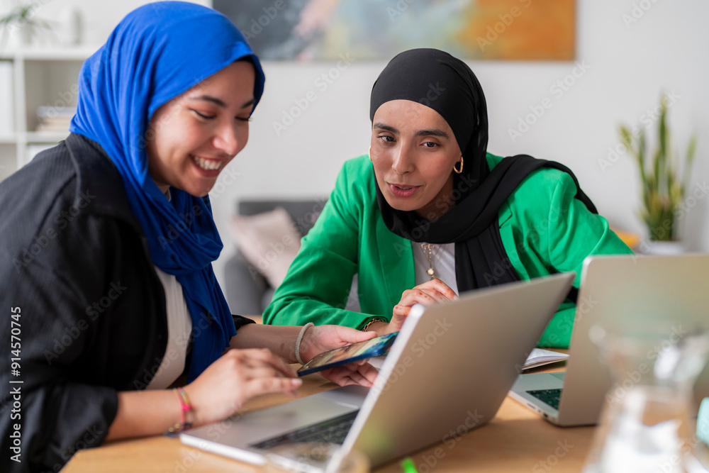Two women in hijabs focus intently on their laptops while discussing work at a desk. Their expressions show determination and concentration in a collaborative office environment. Two women in hijabs focus intently on their laptops while discussing work at a desk. Their expressions show determination and concentration in a collaborative office environment.