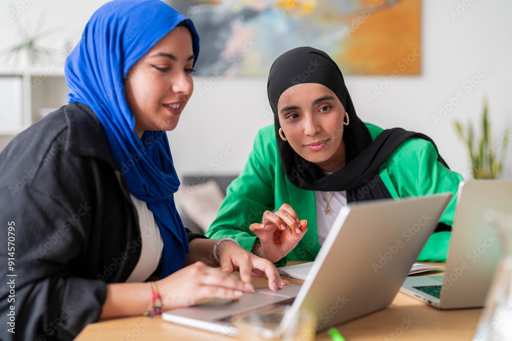 Two women in hijabs focus intently on their laptops while discussing work at a desk. Their expressions show determination and concentration in a collaborative office environment. Two women in hijabs focus intently on their laptops while discussing work at a desk. Their expressions show determination and concentration in a collaborative office environment.