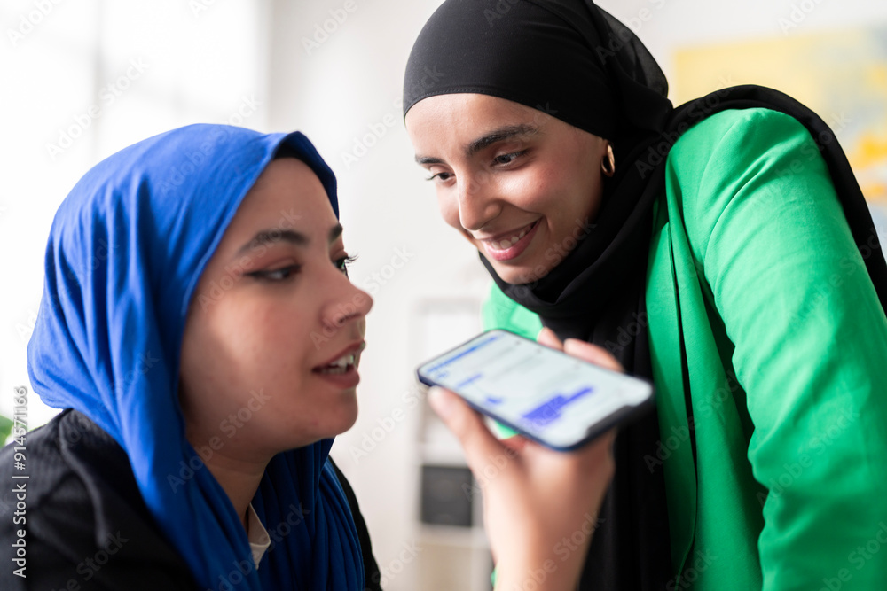 Two women in hijabs recording a voice message on mobile phone in a bright office. Their joyful expressions add to the friendly and positive work environment. Two women in hijabs recording a voice message on mobile phone in a bright office. Their joyful expressions add to the friendly and positive work environment.