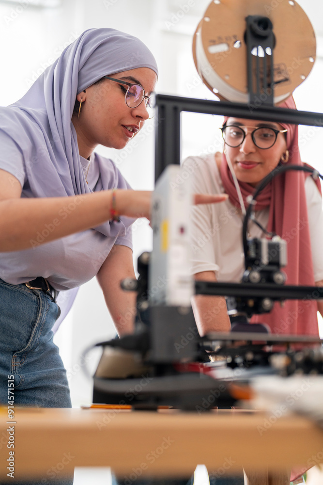 Two women in hijabs collaborate on setting up a 3D printer, carefully adjusting and calibrating the machine. Their teamwork emphasizes precision and technical skills in engineering. Two women in hijabs collaborate on setting up a 3D printer, carefully adjusting and calibrating the machine. Their teamwork emphasizes precision and technical skills in engineering.