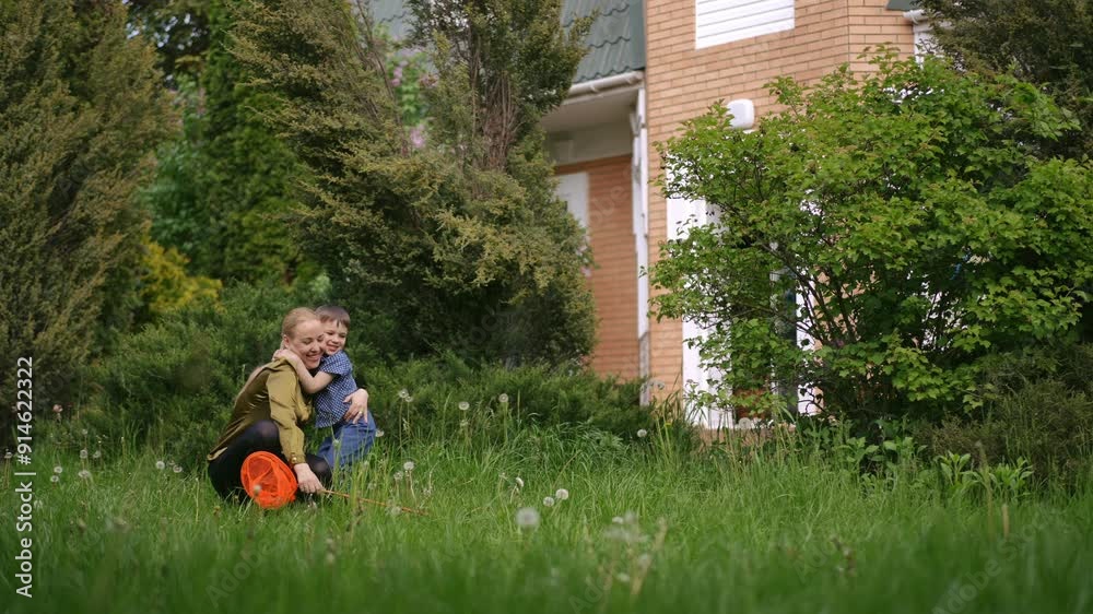 Slow motion. The boy sprints toward his mother, who is squatting outdoors with a net in her hand in the grass near the house. A little son tenderly hugs his mother Slow motion. The boy sprints toward his mother, who is squatting outdoors with a net in her hand in the grass near the house. A little son tenderly hugs his mother