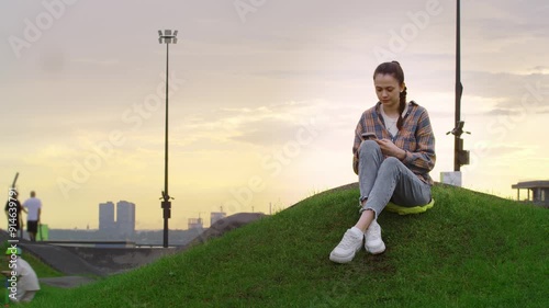 Young woman smiled rather flirtatiously while texting with boyfriend in a city park at sunset. photo