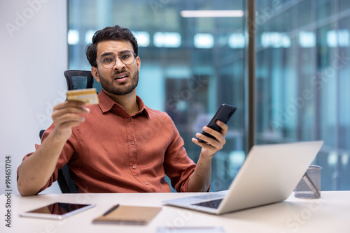 Frustrated man holding bank card and phone at office desk with laptop expressing concern about online fraud. Concept of financial problem, fraud, and technology misuse