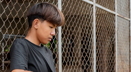 Asian teenboy in a black shirt sits pressed against a metal fence panel in a juvenile detention facility lonely, awaiting further release, depression,freedom and detention of young people concept. photo