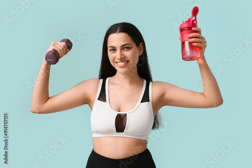 Smiling young woman in sportswear holding dumbbell and shaker bottle, fitness...