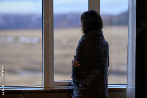Woman wrapped in blanket looking out window at overcast landscape