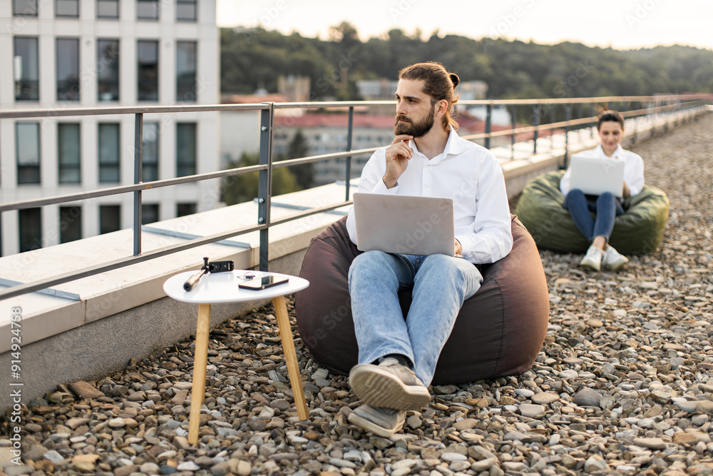 Two people working on laptops in bean bag chairs on rooftop. Relaxed work environment with cityscape in background. Man in foreground looks thoughtful.