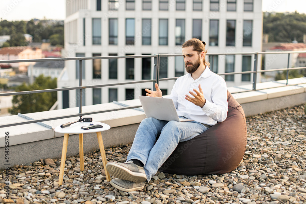 Man working remotely on rooftop while engaging in video call on laptop. Casual work setup with bean bag chair and small table with electronics in outdoor setting. Man working remotely on rooftop while engaging in video call on laptop. Casual work setup with bean bag chair and small table with electronics in outdoor setting.