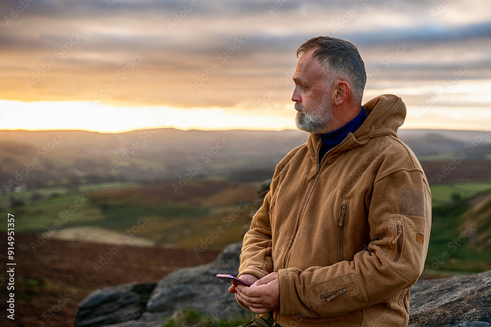 Man With Beard Stands on Rocky Overlook, Holding Phone, as Sun Sets Over Scenic Landscape in Countryside Man With Beard Stands on Rocky Overlook, Holding Phone, as Sun Sets Over Scenic Landscape in Countryside