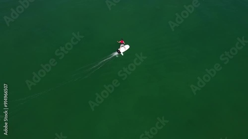 A man in a red vest and hard hat riding on an electric surfboard aerial view. Electric surfboard with rider moving on the water. Man on electric water scooter movement on water top view. photo