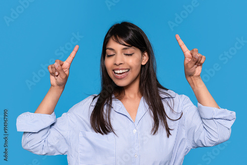 Young Woman Wearing Striped Shirt Celebrating With Her Hands Raised Against Blue Background