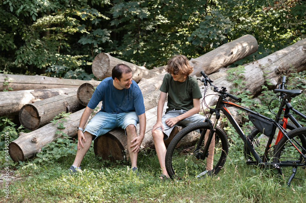 Teenager boy and his father sitting on logs in forest with bicycles, family have a break during cycling outdoor in nature Teenager boy and his father sitting on logs in forest with bicycles, family have a break during cycling outdoor in nature