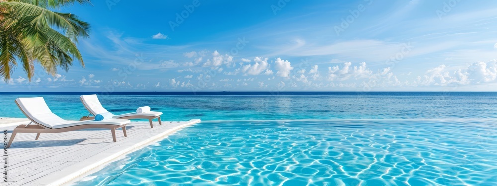 Fototapeta premium A pair of lounge chairs sits at the pool's edge, facing the ocean A palm tree stands prominently in the foreground