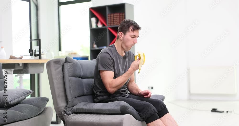 Man sits comfortably on soft sofa and eats ripe banana. Male enjoys natural sweetness of fruit. Guy enjoys healthy snack during break in office hall Man sits comfortably on soft sofa and eats ripe banana. Male enjoys natural sweetness of fruit. Guy enjoys healthy snack during break in office hall