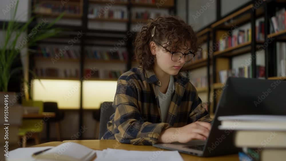 Confident girl with curly hair in a plaid shirt and glasses works at a table using a laptop in a university library Confident girl with curly hair in a plaid shirt and glasses works at a table using a laptop in a university library