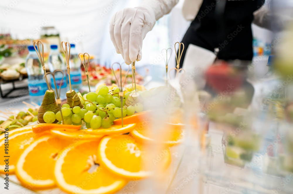 A close-up of a person in white gloves arranging fruit skewers on a platter at a catering event, featuring fresh grapes and orange slices. Perfect for culinary, catering, and event planning themes. A close-up of a person in white gloves arranging fruit skewers on a platter at a catering event, featuring fresh grapes and orange slices. Perfect for culinary, catering, and event planning themes.