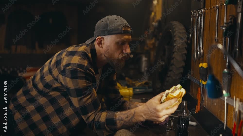 Side view of a mechanic guy in a plaid shirt drinking soda and eating a burger during lunch in a studio workshop