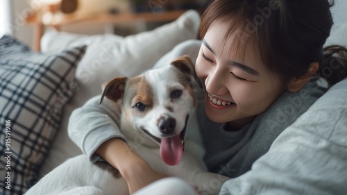 Asian young happy cheerful female owner sitting smiling on cozy sofa couch playing hugging cuddling with best friend small companion dog white short hair parson jack russell terrier in living room.