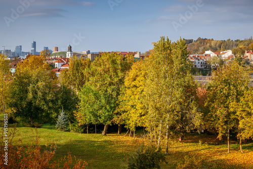 A sweeping autumn view of Vilnius, Lithuania, showcasing golden and orange foliage in the foreground, with urban structures and a towering skyscraper in the background