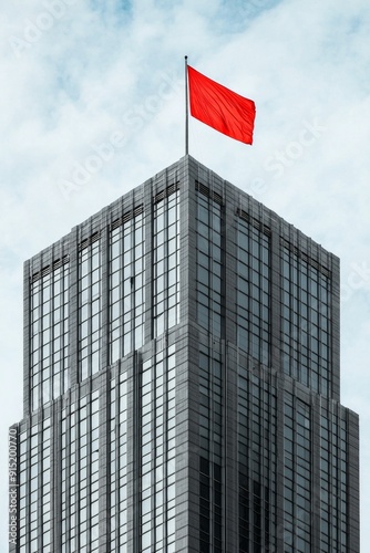 A modern grey skyscraper with clean lines and large reflective windows is punctuated by a single, vividly colored red flag at the top, creating a striking focal point. 