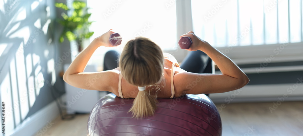 Fototapeta premium A woman lies on a burgundy ball with dumbbells in her hands, rear view, sunlight. Fitness classes at home, aerobics
