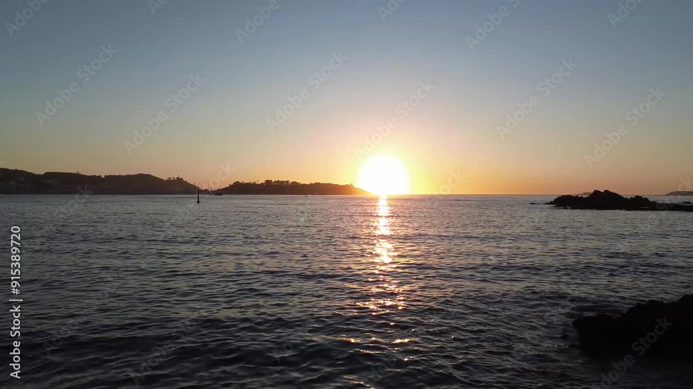 Camera glides from Monte Lourido over calm waters at sunset, capturing the silhouette of the Parador de Baiona against the orange sky. The sun sets beside Baiona as it reflects on the tranquil sea. Camera glides from Monte Lourido over calm waters at sunset, capturing the silhouette of the Parador de Baiona against the orange sky. The sun sets beside Baiona as it reflects on the tranquil sea.