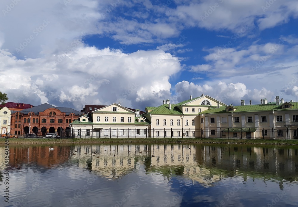A scenic view of historic buildings with green roofs and classical architecture, beautifully reflected in a calm pond. The vivid blue sky with dramatic clouds adds contrast to the serene waters