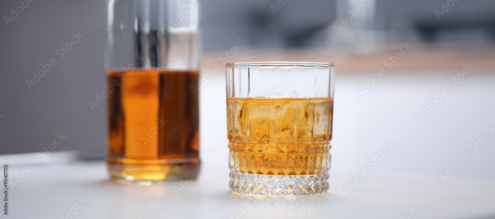 Brown alcoholic cocktail with ice in a wide glass, close-up. Incomplete whiskey bottle and glass on gray table background Brown alcoholic cocktail with ice in a wide glass, close-up. Incomplete whiskey bottle and glass on gray table background