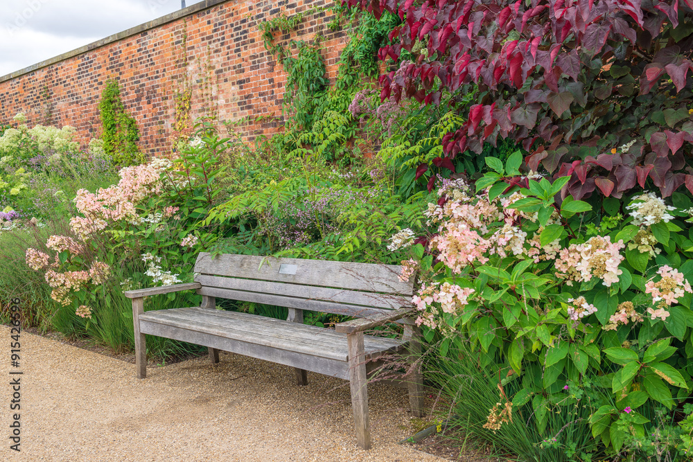 Large wooden bench surrounded by flowering plants in a walled garden. Large wooden bench surrounded by flowering plants in a walled garden.
