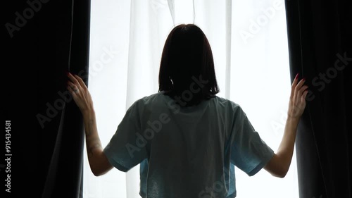 A young woman in a t-shirt opens the dark curtains in the room