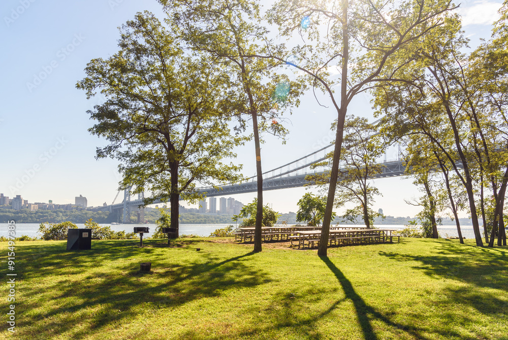 Obraz premium Picninc tables and barbeque grills on grass among trees in a riverbank park on a sunny autumn day. A lage supension road bridge is in background.