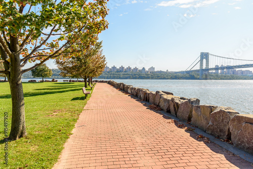 Empty riverbank footpath lined with trees and benches on a sunny autumn day. Apartment blocks and a suspension bridge are visible in background . photo