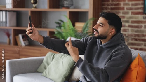 Profile of brunette man with beard and moustache recording video on frontal camera and joking while sitting at home. Enthusiastic blogger promoting infoproducts and showing thumb up with smile. photo