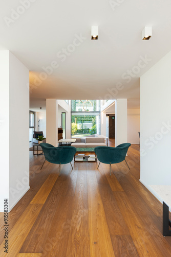 Modern living room with two turquoise armchairs, a coffee table and a sofa with cushions. Large windows overlooking the garden