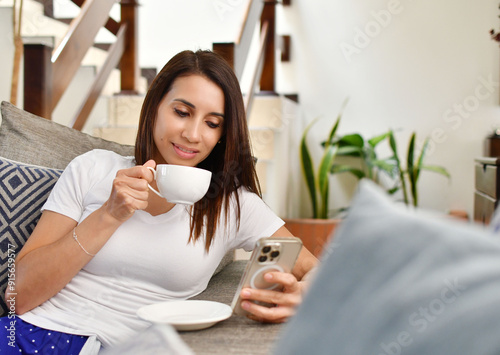 Φωτογραφία Mujer joven latina sentada en la sala de su casa revisando mensajes en su teléfono móvil mientras disfruta de una taza de café por la mañana